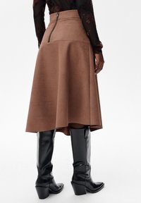 Kobieta w brązowej zamszowej spódnicy midi z zapięciem z tyłu oraz czarnych skórzanych kozakach do kolan, stojąca na białym tle.