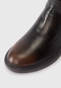 Közelkép barna bőr csizmáról, sima felszínnel és fekete talppal, fehér háttér előtt.