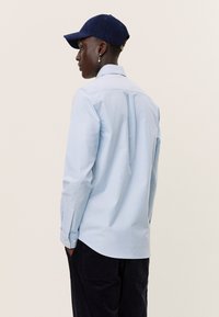 Light blue button-up shirt with long sleeves, tailored fit, back pleat detail, paired with a navy cap, worn with dark corduroy pants.