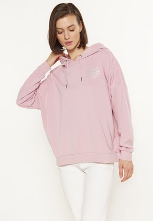 Vrouw draagt een losse roze hoodie met een klein bloemenmotief en witte broek, poseert met één hand die haar nek aanraakt.