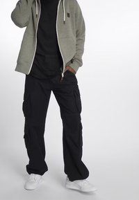 Šedá mikina s kapucí na zip, pod ní černé tričko, černé cargo kalhoty se bočními kapsami a bílé tenisky.