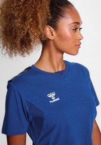 Blå atletisk t-shirt med korte ærmer, V-hals design, mesh-paneler på siden og et hvidt "hummel" logo på brystet.
