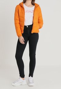 Orange pufferväst med huva, dragkedja och vadderad design. Bärs över en vit T-shirt och svarta skinny jeans, kompletterat med vita sneakers.