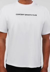 Hvit bomulls t-skjorte med korte ermer, med et svart trykt "CONCEPT SPORTS CLUB"-logo over brystet. Glatt tekstur.