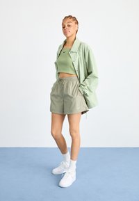 Svetlozelená oversized bunda, zodpovedajúci skrátený top a khaki kraťasy. Biele tenisky Nike a členkové ponožky. Jednoduchý, športový dizajn.