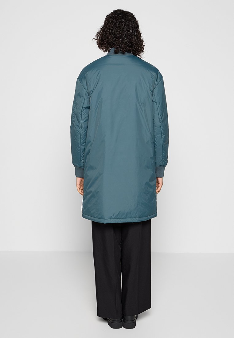 Langer türkisfarbener Steppmantel mit Kragen, gestepptem Muster und gerippten Bündchen; hat einen geraden Schnitt und reicht bis unter das Knie.