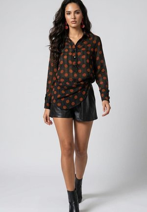 Mujer que lleva una camisa negra con grandes lunares marrones, pantalones cortos de cuero negro, botas negras hasta el tobillo y pendientes rojos caminando hacia adelante.