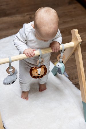 Holzspielbogen für Babys mit drei weichen Spielzeugtieren: einem grauen Koala, einem braunen Löwen und einem grünen Vogel, die alle an blauen Gurten aufgehängt sind. Weicher weißer Teppich darunter.