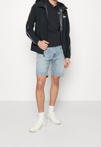 Černá bunda na zip s kapucí, světle šedý polo a modré džínové šortky. Bílé tenisky s několika pásky dotvářejí vzhled.