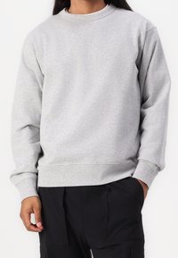 Šedá mikina vyrobená z bavlněné směsi, s kulatým výstřihem, dlouhými rukávy a ribbovanými manžetami a lemem. Nosit s černými kalhotami.