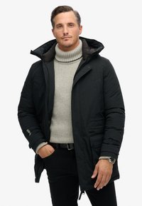 Čierna izolovaná parka s odnímateľnou kapucňou, prednými vreckami a šedým rolákom. Textúra sa zdá byť hladká, dizajn je ležérny.