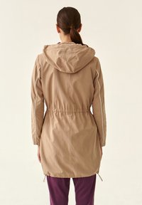 Giacca lunga beige con cappuccio, vita elastica e piping bianco sui lati. Realizzata in tessuto morbido, arriva a metà coscia e presenta maniche lunghe.