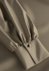 Beiger Langarmhemdärmel mit gerafftem Stoff und einem glänzenden runden goldenen Manschettenknopf auf einem passenden beigen Hintergrund.