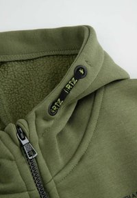 Zielona bluza z kapturem zapinana na zamek, z podszewką z polarowego materiału, zdobiona akcentami z troczkami oraz czarnym zamkiem błyskawicznym. Posiada gładki materiał zewnętrzny oraz wzmocnione przeszycia.