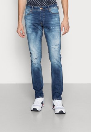 Mann trägt ausgewaschene blaue zerrissene Jeans und weiße Sneakers, steht vor einem schlichten hellen Hintergrund.