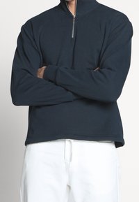 Marine blauwe pullover sweatshirt met een hoge kraag en een kwart rits. Het heeft lange mouwen en een ontspannen pasvorm, gecombineerd met witte broek.