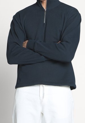 Marine blauwe pullover sweatshirt met een hoge kraag en een kwart rits. Het heeft lange mouwen en een ontspannen pasvorm, gecombineerd met witte broek.
