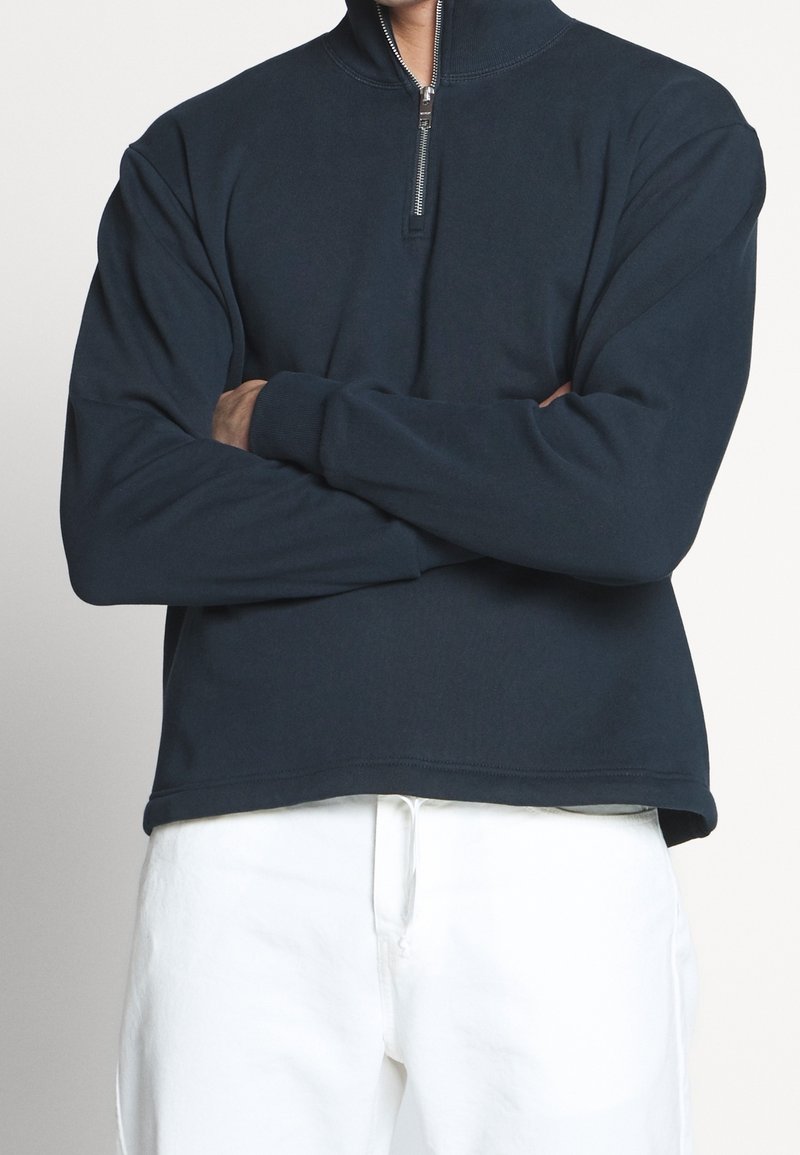 Marine blauwe pullover sweatshirt met een hoge kraag en een kwart rits. Het heeft lange mouwen en een ontspannen pasvorm, gecombineerd met witte broek.