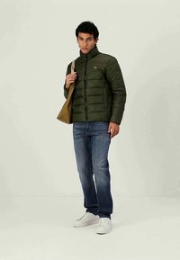 Mörkgrön puffjacka med dragkedja och sydda paneler, kombinerad med blå jeans och vita sneakers; modellen håller en brun axelväska.