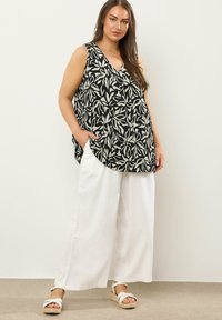 Mujer con blusa sin mangas de patrón floral en blanco y negro, pantalones anchos blancos y sandalias de cuña blancas, posando contra un fondo liso.