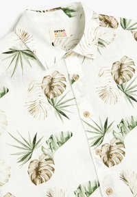 Witte kortarmige blouse met een tropisch bladenpatroon in groen en bruin, met een kraag en lichte houten knopen.