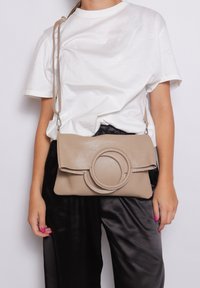 Borsa a tracolla in pelle beige con un accento circolare, dotata di chiusura a patta e tracolla regolabile. Superficie testurizzata con una finitura liscia.
