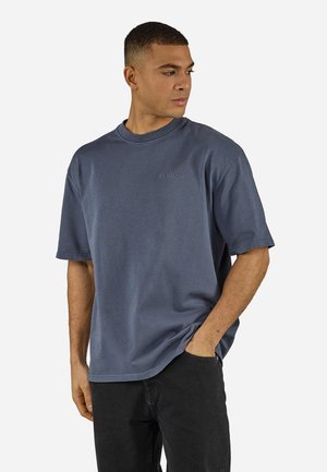 Mann iført en tettsittende mørkeblå t-skjorte med rund hals og svarte bukser, stående med den ene hånden i lommen, og ser mot høyre.