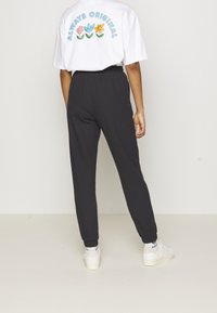 Henkilö, joka käyttää valkoisia lenkkareita, mustia ribattuja joogahousuja ja valkoista T-paitaa, jossa on sininen "Always Original" -teksti ja värikkäitä kukkia takana.