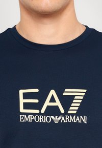 Tamsiai mėlynas apvalaus kaklo marškinėliai, pagaminti iš medvilnės, su kreminės spalvos tekstūriniu logotipu, kuriame užrašyta "EA7 EMPORIO ARMANI" per krūtinę.