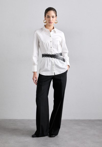 White button-up shirt paired with high-waisted black trousers. Black latex belt with circular cutouts. Large gold hoop earrings.