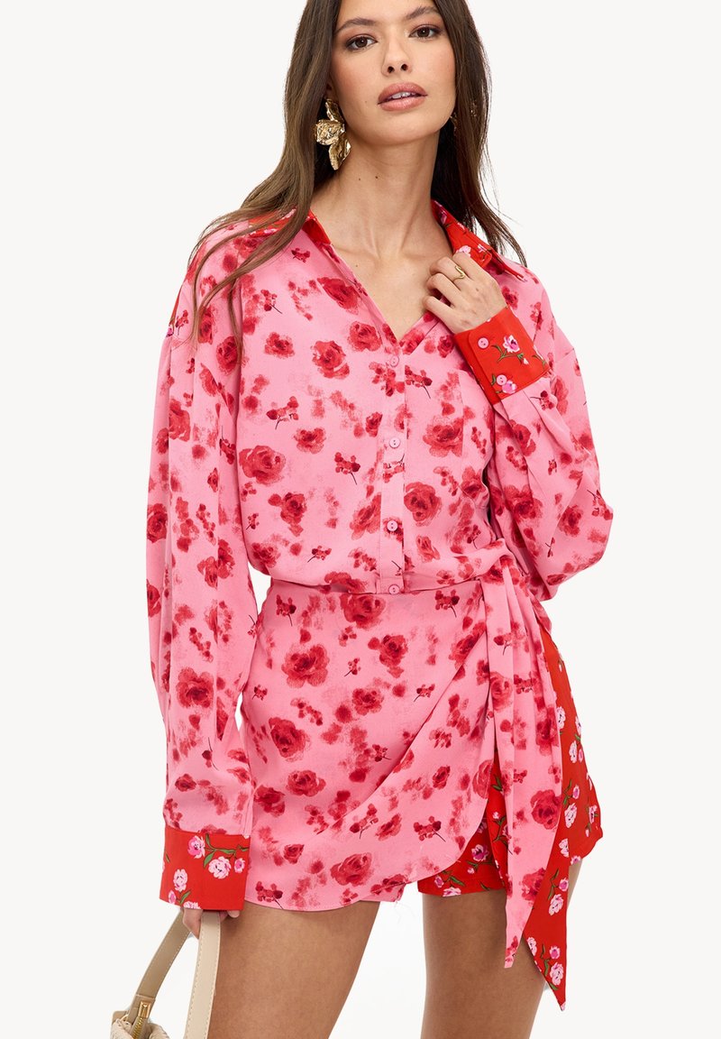 Femme portant un chemisier boutonné à fleurs roses et rouges avec une jupe portefeuille assortie, tenant un sac à main beige, avec des boucles d'oreilles en forme de fleurs dorées.