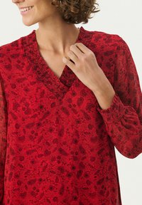 Rote Bluse mit floralem Muster, V-Ausschnitt mit gerafftem Detail und langen, leicht puffigen Ärmeln. Hergestellt aus leichtem Stoff.