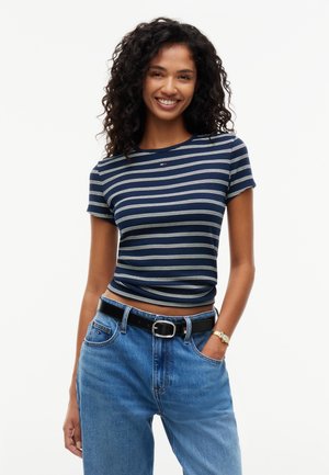 Lächelnde Frau mit lockigem Haar trägt ein marineblau gestreiftes Kurzarmhemd, blaue Jeans mit schwarzem Gürtel und eine goldene Armbanduhr, steht mit einer Hand in der Tasche.