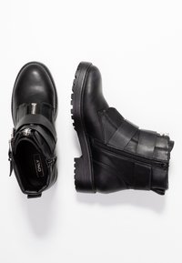 Un par de botas de cuero negro hasta el tobillo con correas y cremallera frontal, una bota en posición vertical y la otra de lado sobre un fondo blanco.