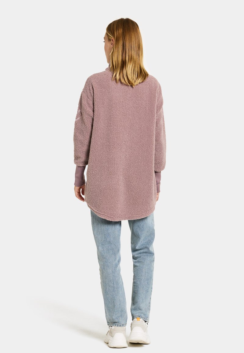 Übergroßer Fleecetop in hellem Mauve, mit langen Ärmeln und abgerundetem Saum. Kombiniert mit hellblauen Jeans und weißen Sneakers.