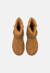 Botas de tobillo de ante color arena con forro de piel sintética, que presentan una correa decorativa con tachuelas circulares alrededor del tobillo. Suelas planas con detalles de costura.
