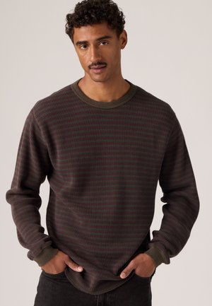 Homme aux cheveux bouclés et à la moustache portant un pull à manches longues rayé vert foncé et bordeaux, les mains dans les poches, debout devant un fond uni.
