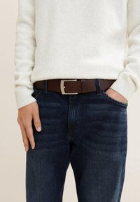 Ceinture en cuir marron avec une boucle en métal argenté, portée sur un jean en denim bleu foncé, associée à un pull en tricot texturé de couleur claire.