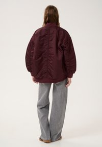 Burgundy puffer jacket with a relaxed fit, ribbed cuffs, and a smooth, shiny texture. Paired with wide-leg gray denim pants.