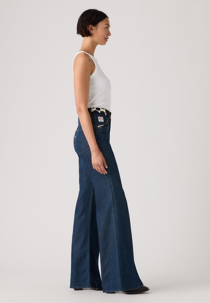 Høj talje mørkeblå flare-jeans med sideskår, lavet af denim, med et mønstret taljebånd og lynlås lukning. Glat tekstur.