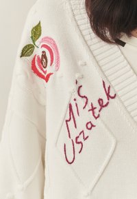 Biały dzianinowy kardigan z teksturowanymi wzorami, haftem róży w odcieniach różu i czerwieni oraz odręcznym napisem "Miś Uszatek" w kolorze fioletowym.