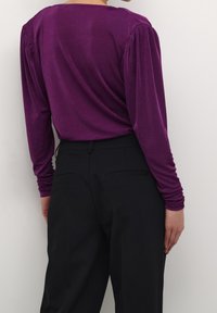 Haut violet à manches longues avec épaules froncées, texture douce et coupe décontractée, associé à un pantalon noir avec poches arrière.