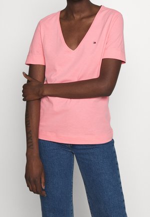 Person wearing a pink V-neck short sleeve shirt with a small logo on chest, blue jeans, and arm crossed holding opposite elbow.