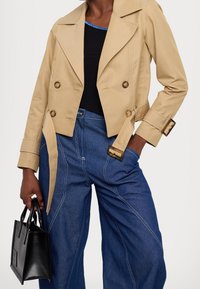 Beige dubbelzijdige jas met gouden knopen, cropped ontwerp en opgerolde mouwen, gedragen over een zwarte top en gecombineerd met brede blauwe denim broeken en een zwarte tas.