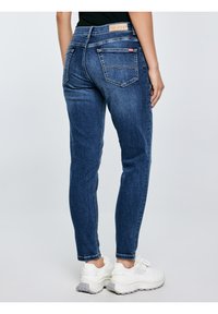 Sötétkék farmernadrágot és fehér tornacipőt viselő személy, aki egy kezét laza tartással oldalán áll, egyszerű világos háttér előtt.