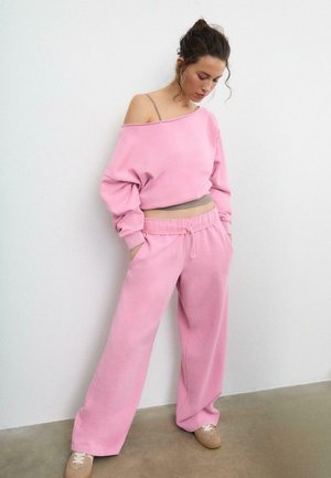Femme portant un sweat-shirt rose ample glissant sur une épaule, pantalon large assorti, baskets beiges, debout contre un mur blanc avec les mains dans les poches.