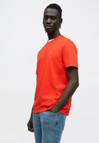 Czerwona koszulka bawełniana z krótkimi rękawami, okrągłym dekoltem i małym detalem z logo. Połączona z jasno-niebieskimi dżinsami. Prosty i casualowy design.