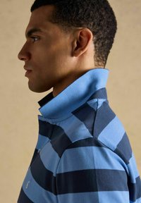 Homme de profil portant un polo à rayures bleues et bleu marine avec le col relevé sur un fond neutre.