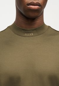 Κοντινό ενός ατόμου που φοράει πουκάμισο ελιάς με ψηλό γιακά, κεντημένο με την ετικέτα "HUGO BOSS" σε νήμα που ταιριάζει στο χρώμα.