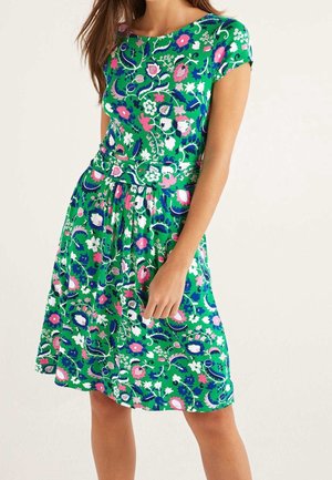 Vrouw draagt een groene jurk tot op de knie met roze, witte en blauwe bloemen- en bladpatronen, korte mouwen en een taille met plooien.